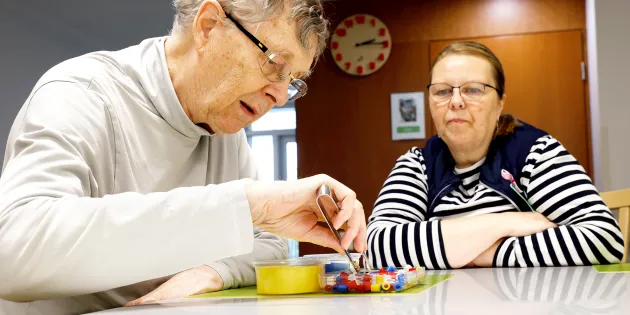 Markku Närhistä auttamassa lähihoitaja Marianne Vierelä. Markku lajittelee pinseteillä pieniä muoviesineitä värin mukaisiin rasioihin.