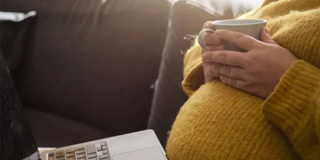 Raskaana oleva nainen siemailee teetä sohvalla ja katsoo kannettavaa.