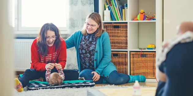 Ida Schroderus loruttelee vauvansa kanssa lattialla. Mirkku Kuivalainen on kaverina vieressä.