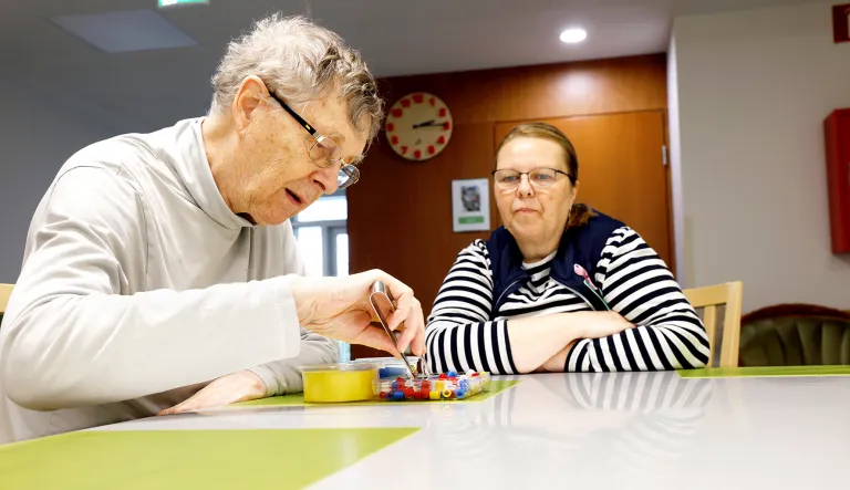 Lähihoitaja Marianne Vierelä istuu vieressä, kun Markku Närhinen laittaa pinseteillä pieniä erivärisiä muovinpalasia erivärisiin kippoihin.